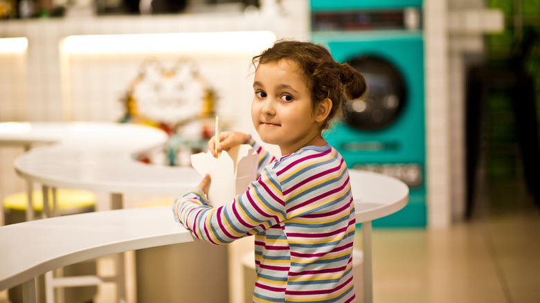 A young girl looks at the camera while she eats noodles in a mall food court