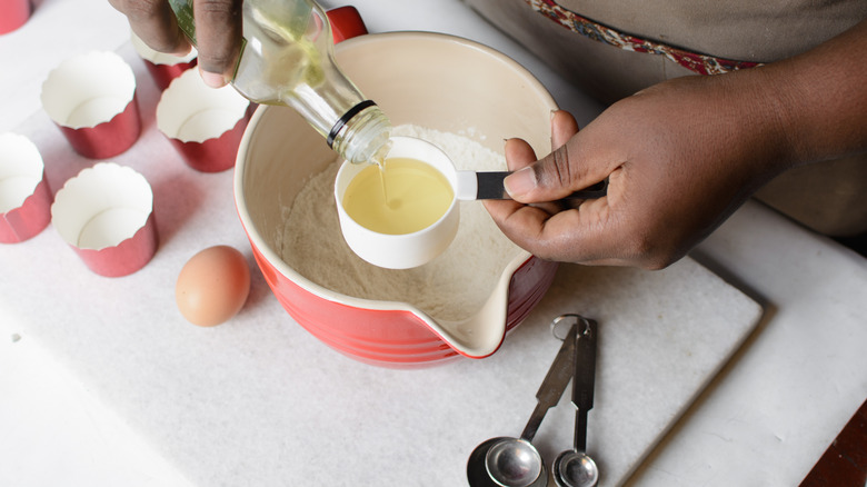 A person carefully pouring oil into a measuring cup over a bowl of flour