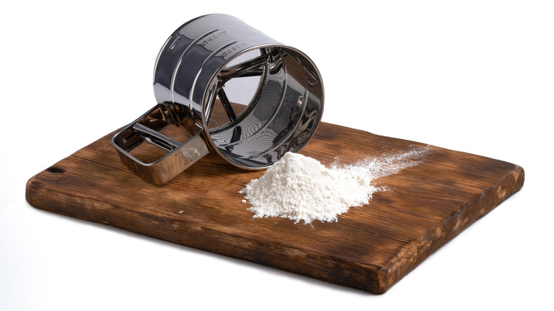 A metal sifter and small pile of white flour on a wooden cutting board