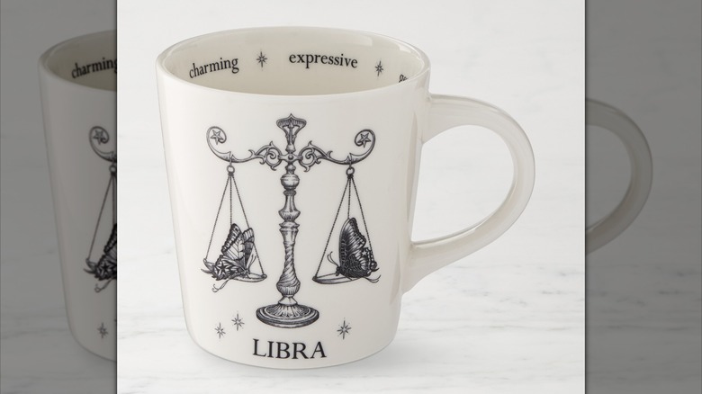 A white coffee mug with an ink drawing of the Libra scales of justice, with a moth on one and a butterfly on the other. Inside of the mug says "charming" and "expressive"
