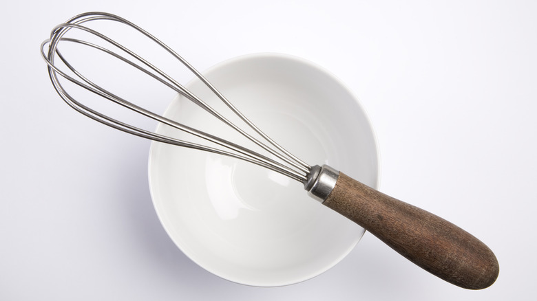 A metal whisk lays atop an empty white mixing  bowl ready for ingredients