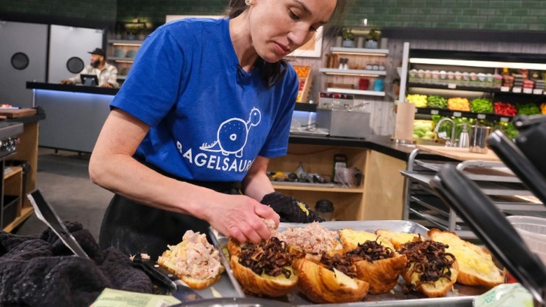 A woman in a blue t-shirt. preparing a tray of bagel BLTs in a restaurant kitchen