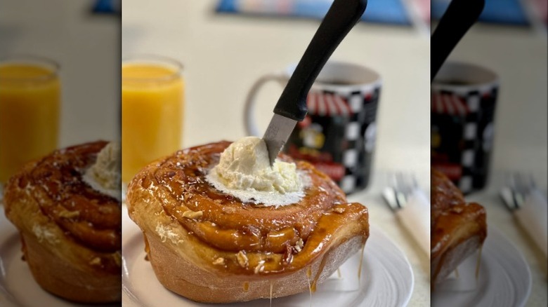A giant pecan roll with cream on top, and a knife ready to cut through, with a coffee mug and a glass of orange juice in the background