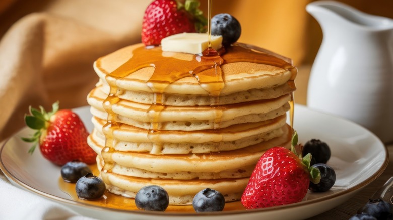 A stack of pancakes with a dab of butter on top, drizzled with  maple syrup, and garnished with strawberries and blueberries