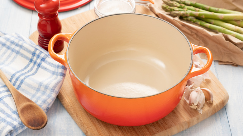 A round orange Dutch oven with the lid removed, on a cutting board surrounded by ingredients