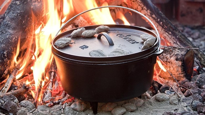 A Lodge Camp Dutch oven in black, sitting inside a campfire with hot coals on its lid
