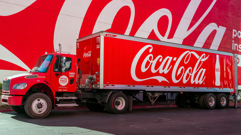 A red Coca-Cola delivery truck covered in logos, parked in front of a mural of a Coca-Cola logo.