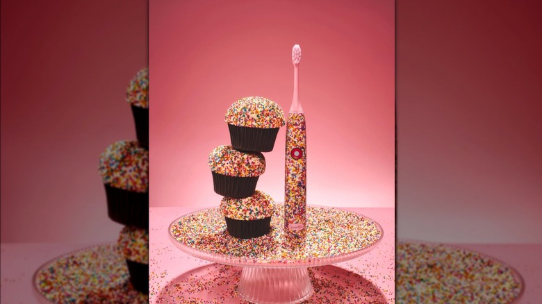 a sprinkles x moon toothbrush next to a stack of cupcakes on a cake stand covered in sprinkles