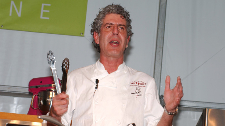 Anthony Bourdain holding a pair of tongs and doing a cooking demonstration