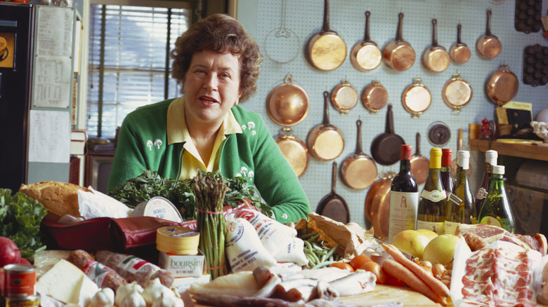 Julia Child sitting beind a counter with piles of produce, meats, and bottles of wine, behind her is a pegboard with hanging copper cookware