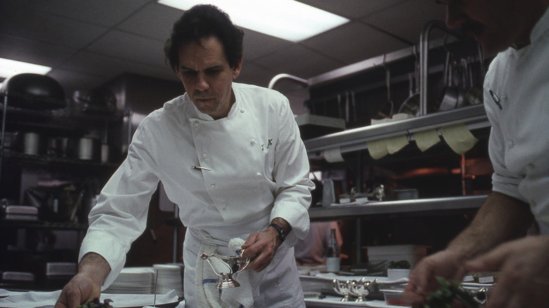 Thomas Keller wearing chef's whites in his kitchen, seasoning a dish while another chef looks on