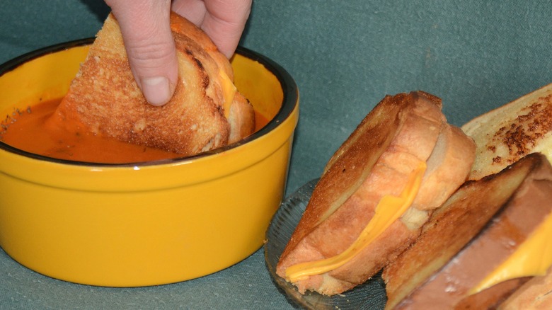 A grilled cheese sandwich is dipped into soup, with a stack of grilled cheese sandwiches sitting next to the soup bowl