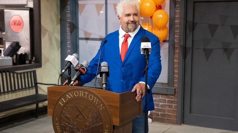 In a still from "Flavortown Food Fight," Guy Fieri stands at a podium as he opens the gates to Flavortown