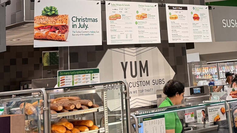 A Publix deli sub counter, signs, and an employee behind the glass display