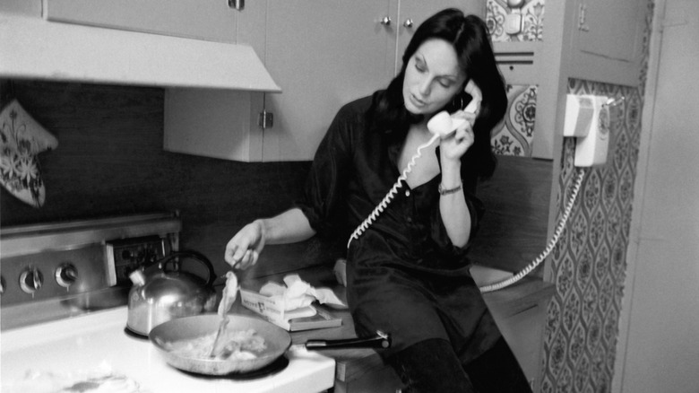Black and white photo of a woman in the kitchen, talking on a corded phone while tending a frying pan