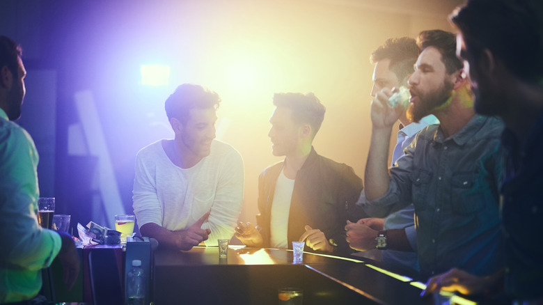 A group of friends chat around a bar, with one of them drinking a shot