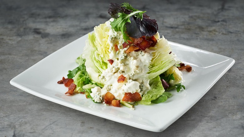 Crisp iceberg wedge salad topped with blue cheese, bacon, and dressing on a white plate.