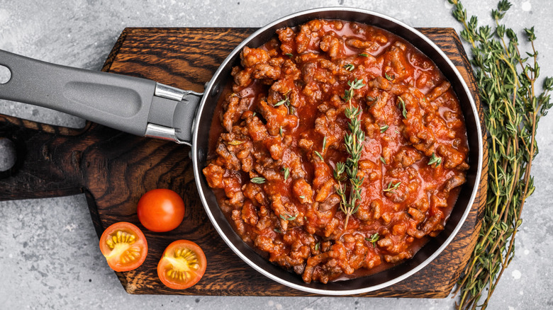 Bolognese sauce in a pan placed on a wooden board with fresh thyme and cherry tomatoes on the side