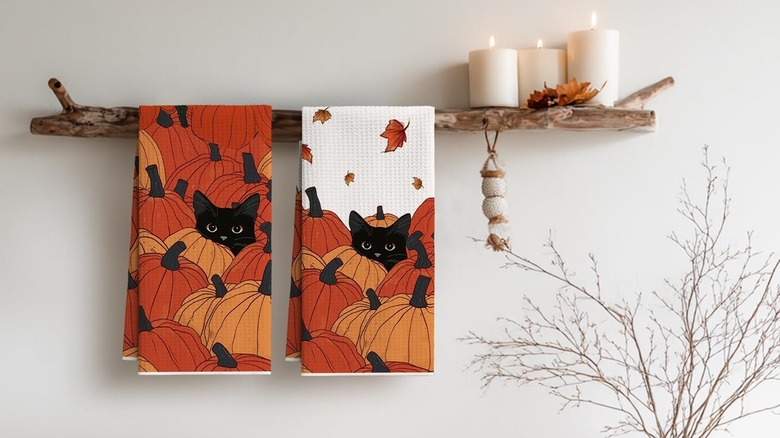 Two tea towels featuring a black cat in a pumpkin patch, hanging from a driftwood shelf