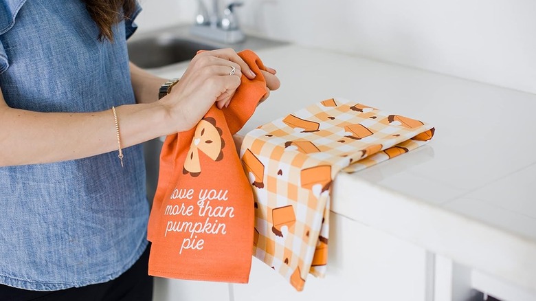 A woman holding an orange tea towel, with a second patterned towel on a kitchen surface in front of her
