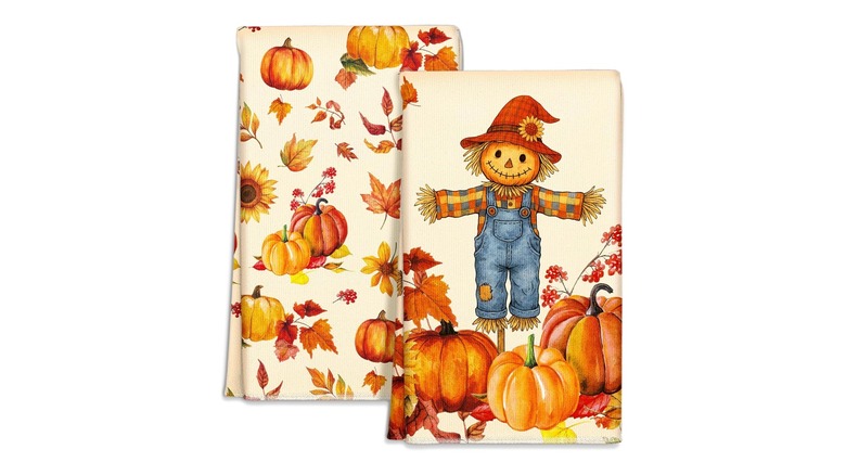 Two folded kitchen towels, one decorated with a scarecrow in a pumpkin field, the other with a pattern of pumpkins and fall leaves