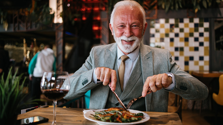 An older businessman sits in a restaurant having lunch, while looking at the camera and smiling