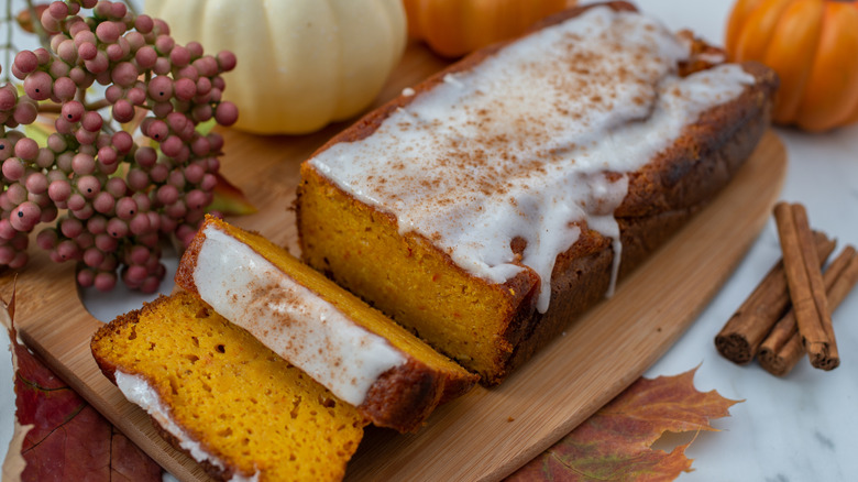 Sliced pumpkin loaf with icing on top on a cutting board with pumpkins and cinnamon sticks around it