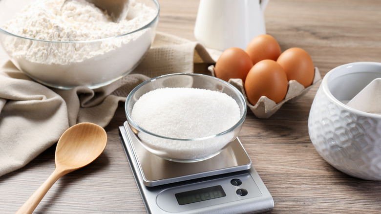 A bowl of sugar on food scale with eggs and flour behind it