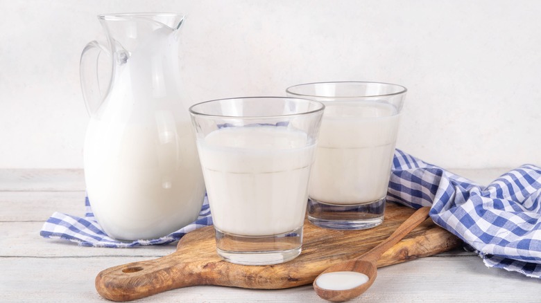 A pitcher and two glasses of buttermilk on a wooden cutting board with a blue and white checkered towel in the background