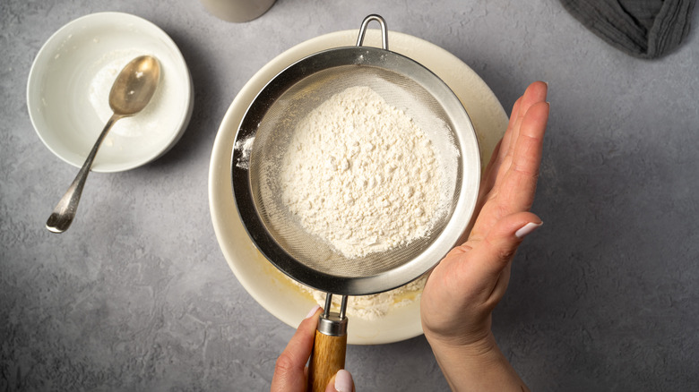 A person sifts flour using a sieve into a mixing bowl
