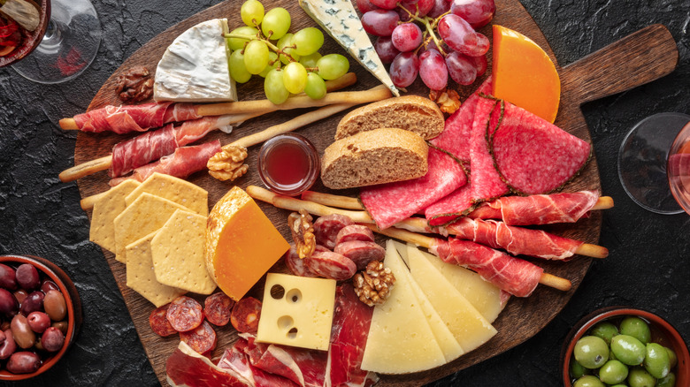 A fruit, meat, and cheese board with bowls of olives on the side