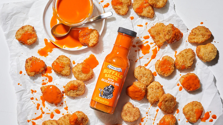An overview of a bottle of Buffalo sauce with chicken nuggets with the sauce drizzled all over the table against a white background.