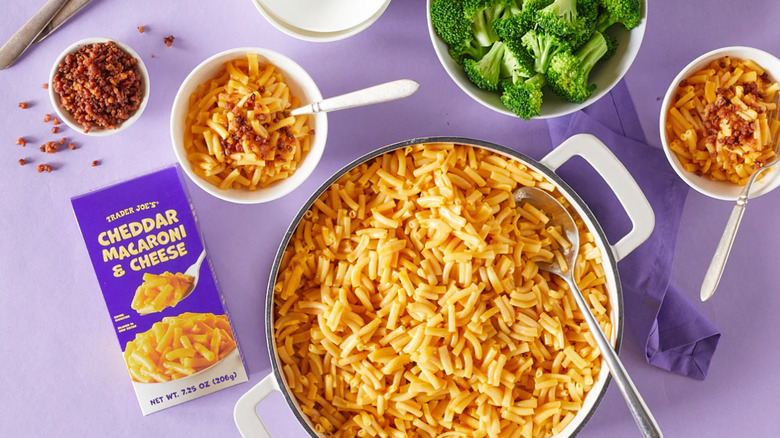 A pot of Mac & Cheese next to some broccoli, two smaller bowls of pasta, and a box of Trader Joe's mac & cheese against a purple table.