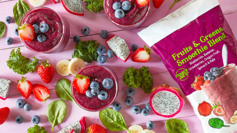 A variety of smoothie glasses, fruits, vegtables, on a pink table next to a package of Trader Joe's smoothie blend