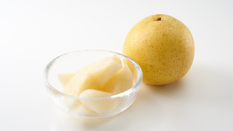a 20th century asian pear next to a glass bowl of sliced pear against a white background
