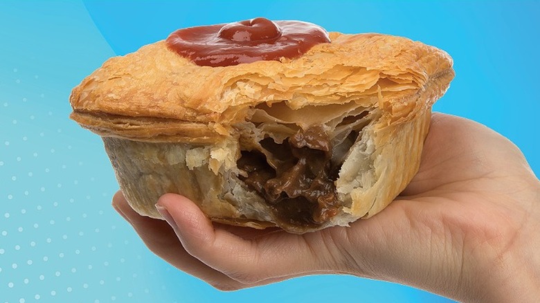 A hand holding an Aussia steak pie with a bite taken out and ketchup on top