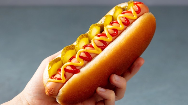 A hand holding a hot dog with pickles, mustard and ketchup on top