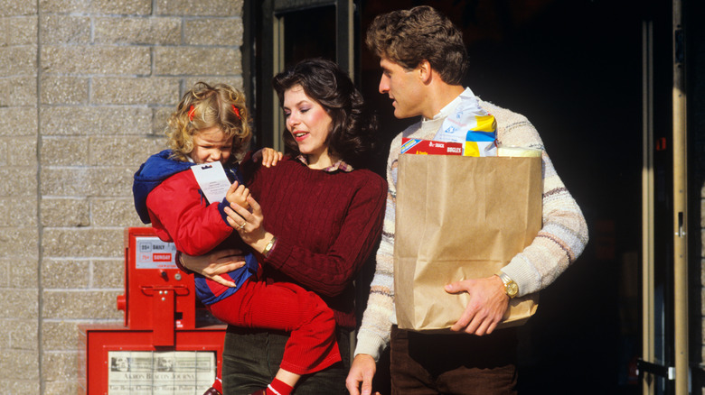 A couple with their child leave a store with a paper bag full of groceries