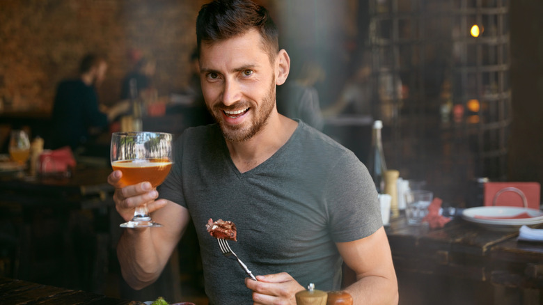 A man eats steak and drinks craft beer in a restaurant, holding up his glass as he smiles at the camera