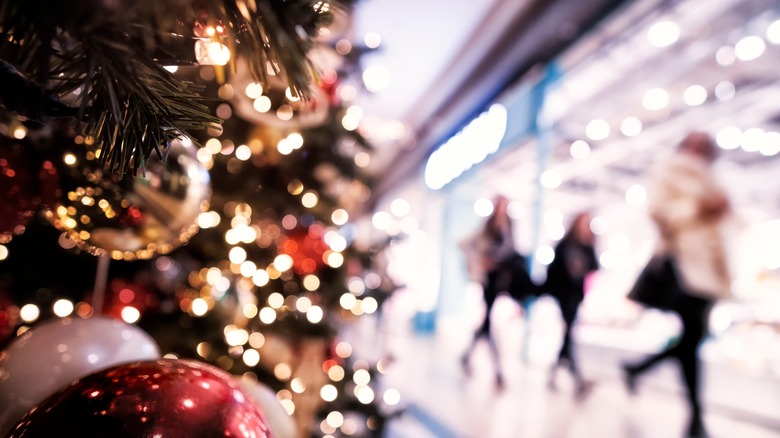 A Christmas tree is framed in the foreground, with blurry shoppers in the background