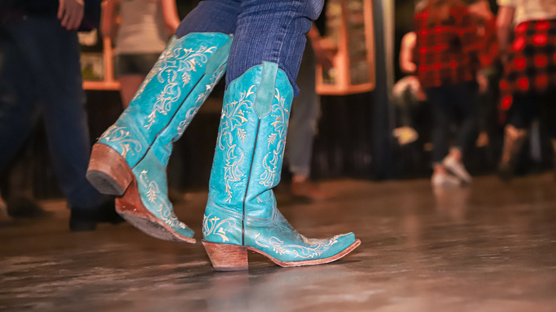 A pair of turquoise boots performs a line dance against a wooden floor