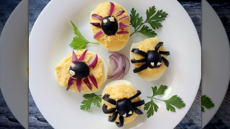 Spider-topped deviled eggs with olive bodies and olive or red onion legs on a white plate with sprigs of herbs and onion slices
