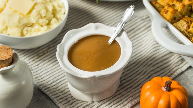 A small white container of gravy sits on a table with other Thanksgiving foods