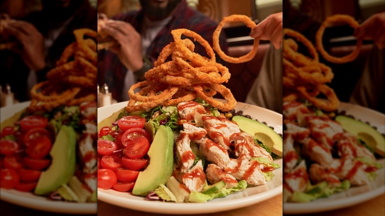 A large salad with onion rings, tomato, avocado, and chicken on a white plate