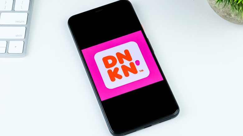 The Dunkin' logo is seen on a smartphone, sitting on a white desk