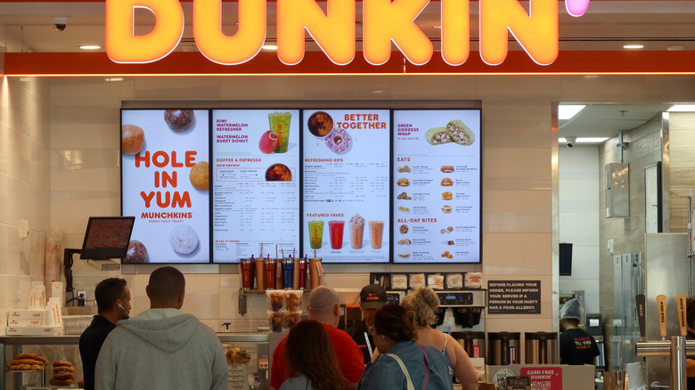 A queue of people stand at a Dunkin' counter, with the menu and the Dunkin' logo seen above