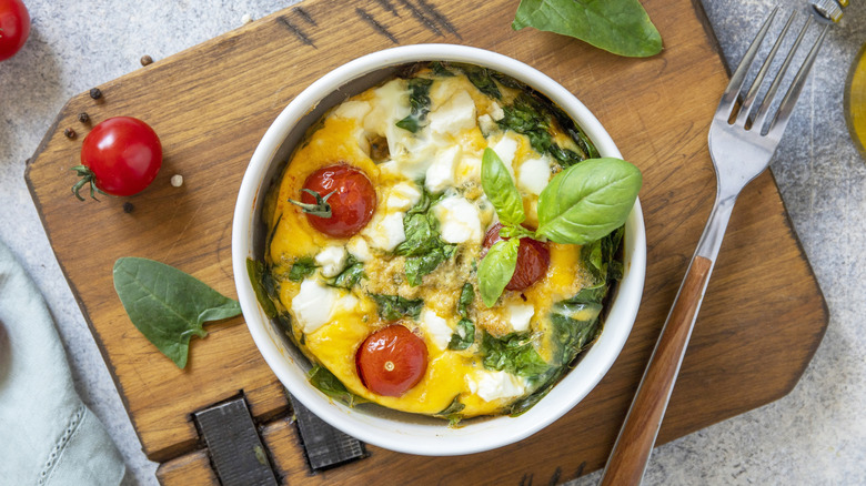 Baked eggs with veggies and cheese in a bowl on a wooden board with a fork on the side