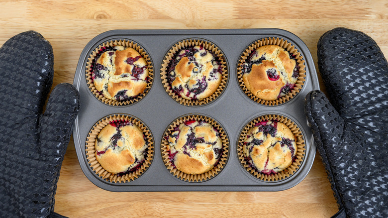 A person wearing oven mitts and holding a muffin pan with baked muffins on a wooden table