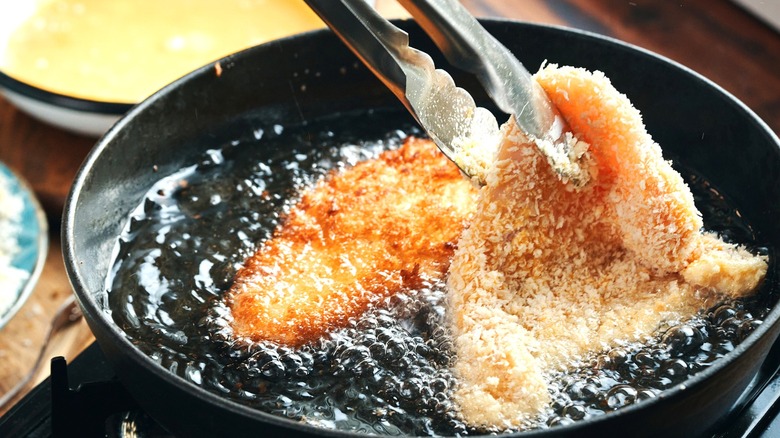 A pair of tongs gripping crispy chicken frying in a pan for Japanese katsu