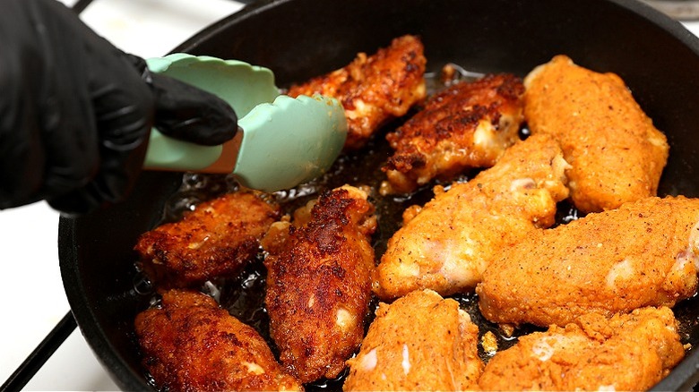 A chef in black gloves picking up chicken wings in a frying pan with tongs